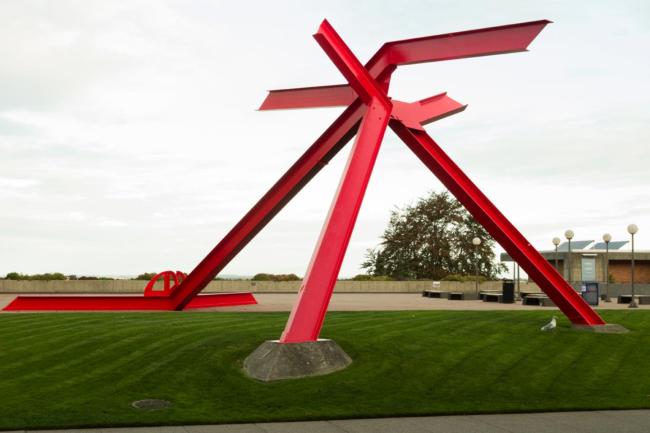 Large metal sculpture straddling a mowed lawn: three legs conjoin in a pyramid shape and then bend outward in four arms pointing different directions.