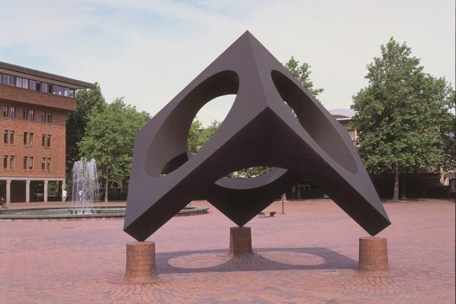 A three sided metal cube with a large hole in each side stands with open corners resting on rounded brick platforms, in Red Square on Western's campus
