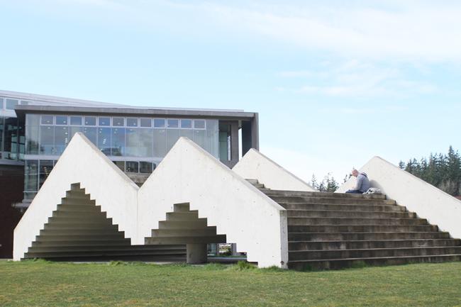 concrete staircase on a lawn leading up and down, form the shape of an M when viewed from the side. A person sits calmly at the top of one of the stair peaks.