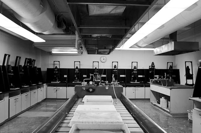 black and white photo of an empty darkroom, showing a film developing station and counters filled with rows of projectors