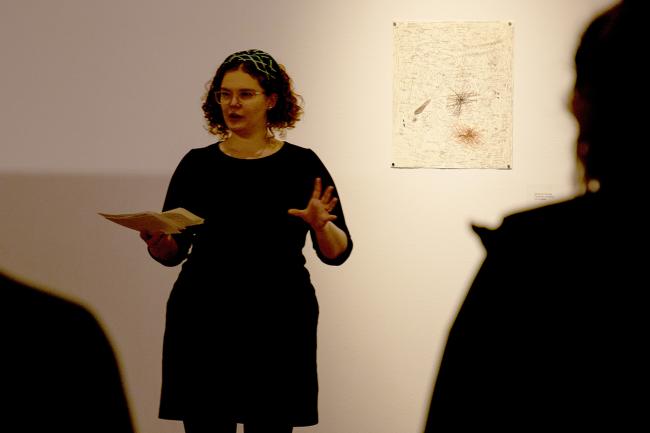 A person stands in front of a piece of artwork hanging in a gallery, presenting to a silhouetted group