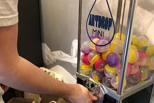 a hand turns the crank of a small vending machine filled with bright eggs containing art