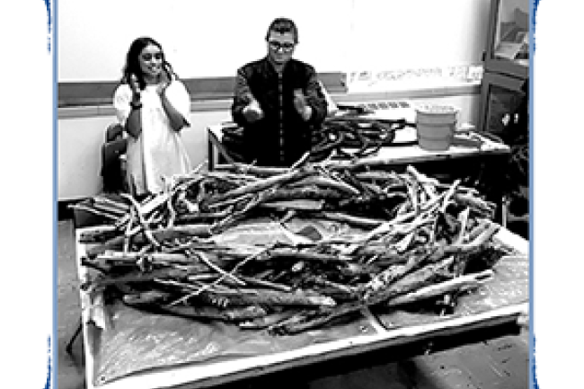two people in a classroom talking over a table which holds a person-sized, bottomless nest made of sticks
