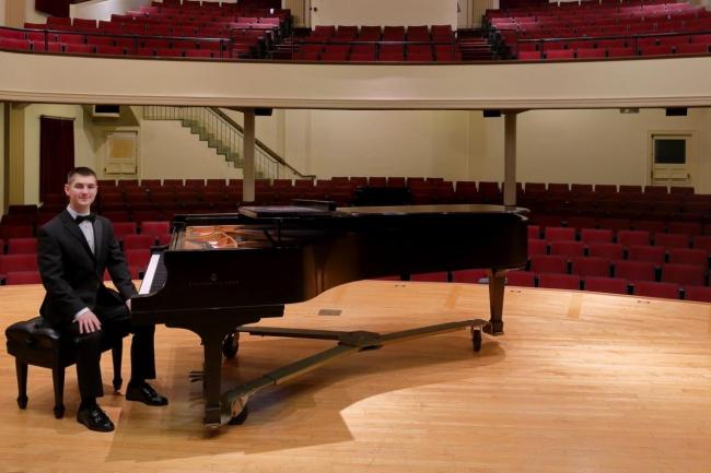 young person seated at a grand piano on a stage in an empty concert hall