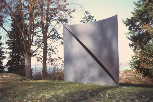 A tall, thin, curved metal square stands on a lawn. A diagonal fold appears to divide it.