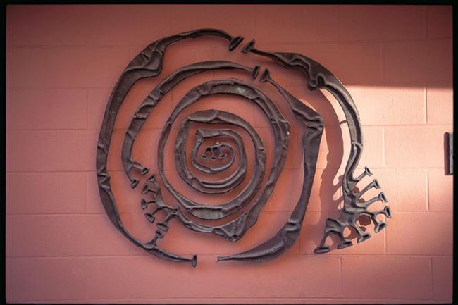 A stylized spiral sculpture made of metal, mounted flat against a brick wall