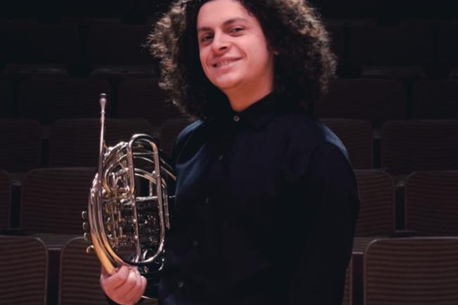 A well dressed person with a long fro smiles and holds a french horn