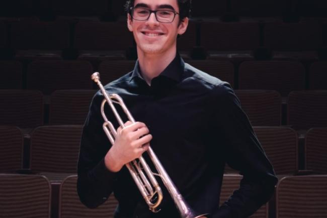 A well dressed person stands mid-chuckle, holding a trumpet to their torso