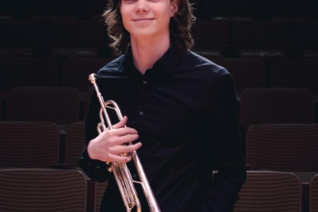 A well dressed person with shoulder length hair smiling, holding a trumpet