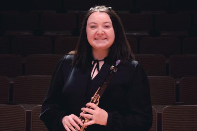 A well dressed person with long hair and a big smile holds a clarinet
