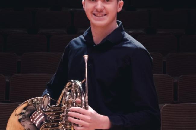 A well dressed person with short hair smiles and stands holding a french horn