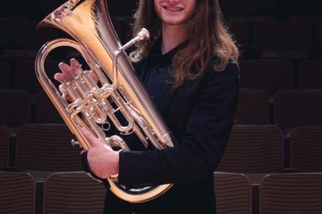 A person dressed formally with long hair smiles and holds up a euphonium