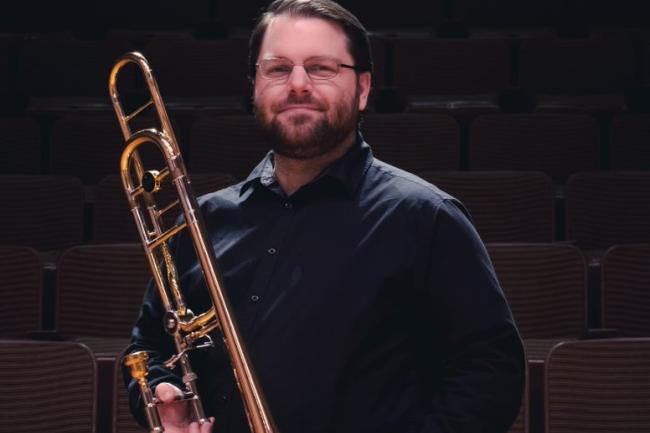 A well dressed person with short hair, glasses and a beard stands proud and calm holding a trombone