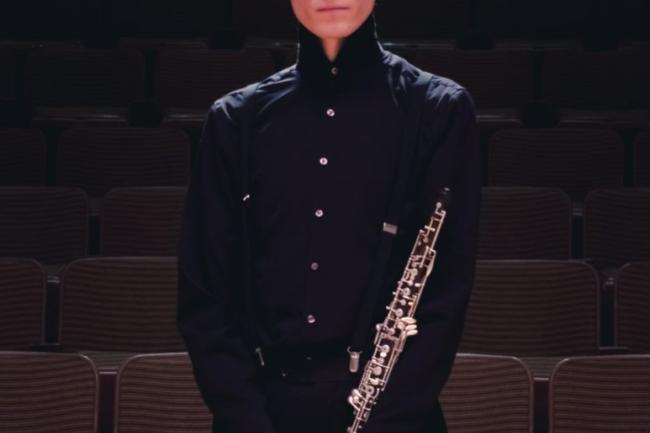 A nicely dressed person stands straight and relaxed holding an oboe in clasped hands, with a somber face