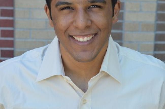 Carlos Cordero in a button up shirt in front of a brick wall, smiling with a squint
