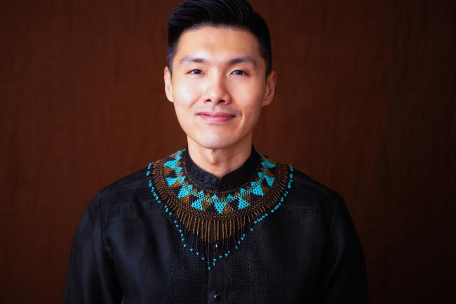 Saunder Choi smiles proudly while wearing a dark top with a large beaded necklace around the collar