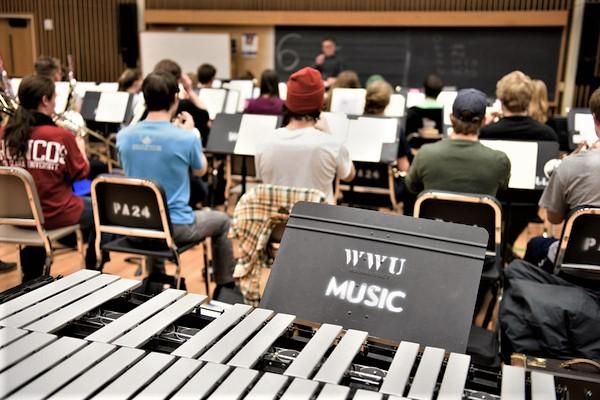 A music stand with "WWU Music" stenciled on it, in front of a class full of students playing instruments
