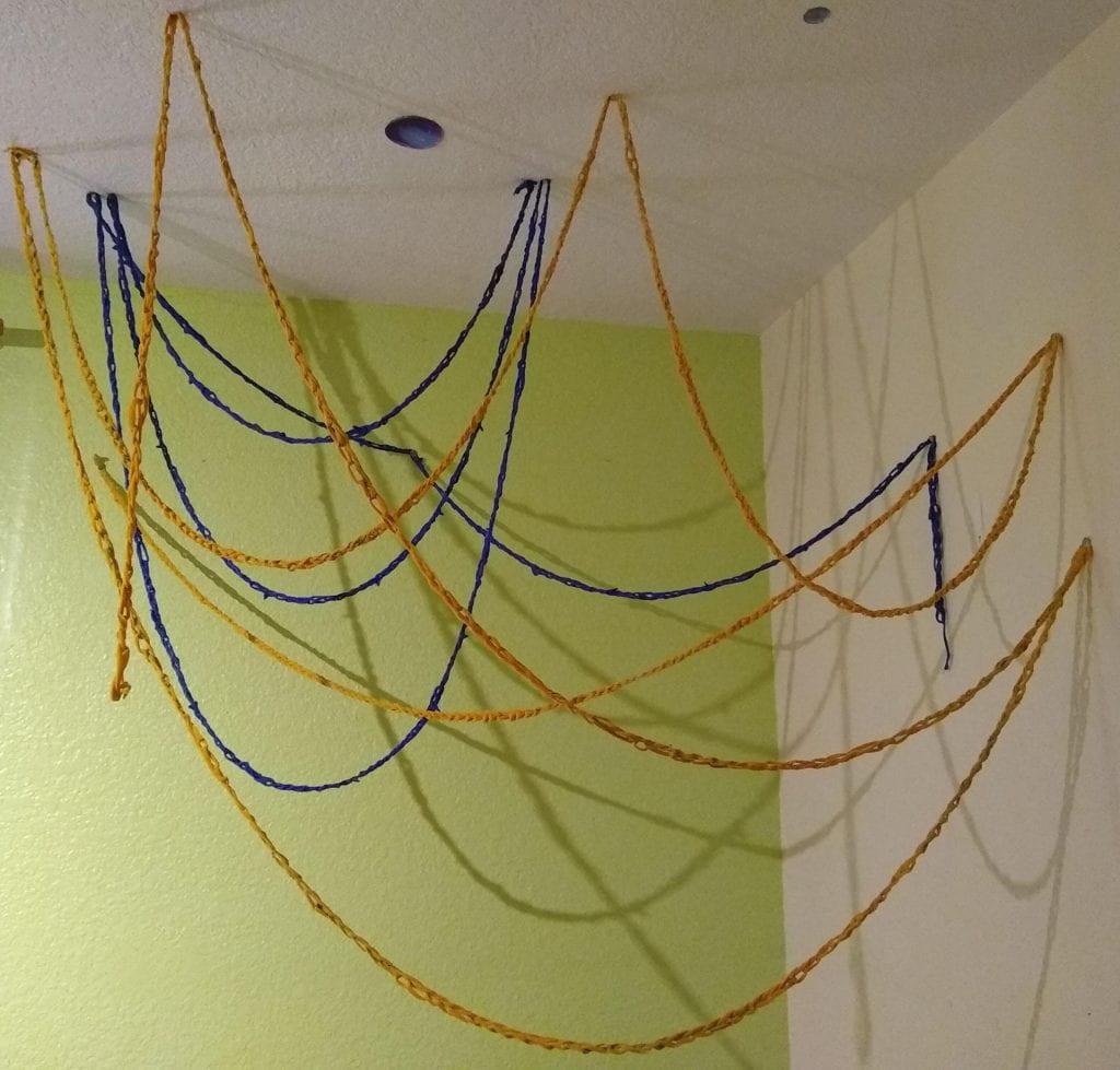 blue and yellow chains made from yarn hang from points on the ceiling and walls in the corner of a room, forming something like a loose net