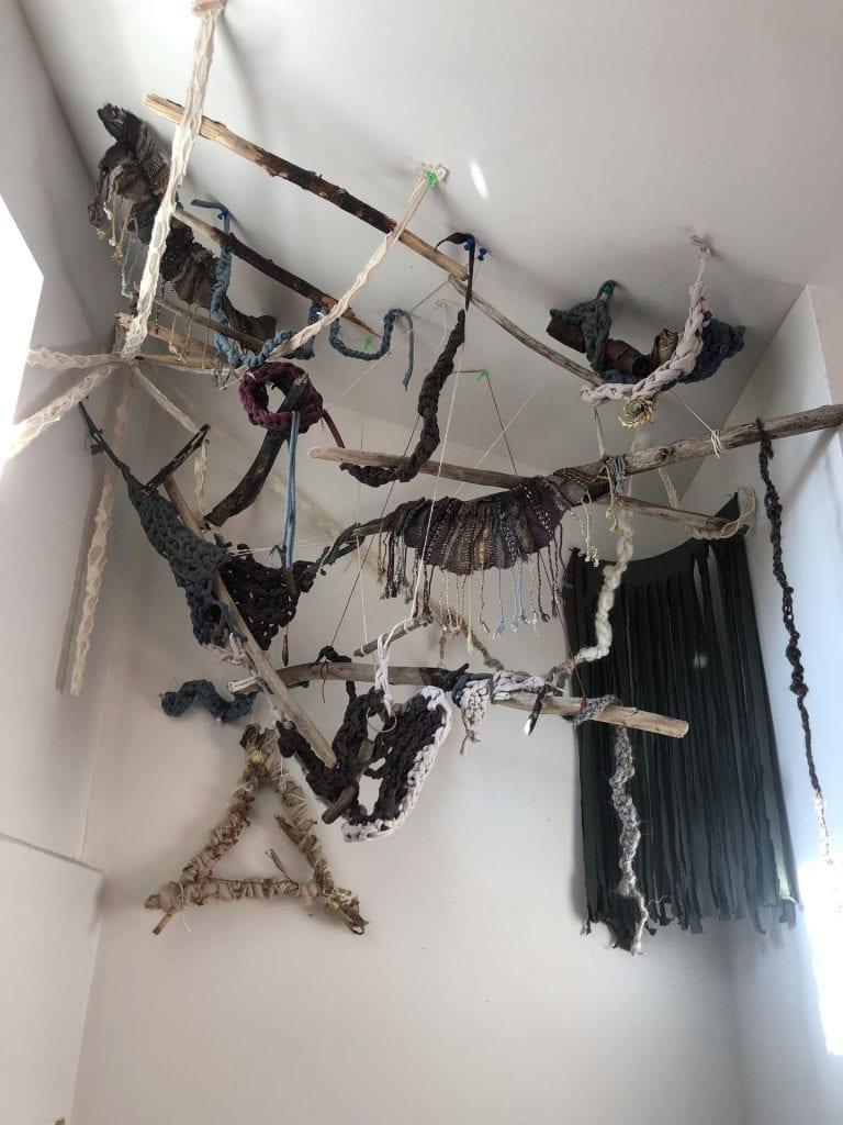 driftwood hangs from the ceiling. Strings, shredded fabric, knitted strands and shapes hang from the driftwood pieces.