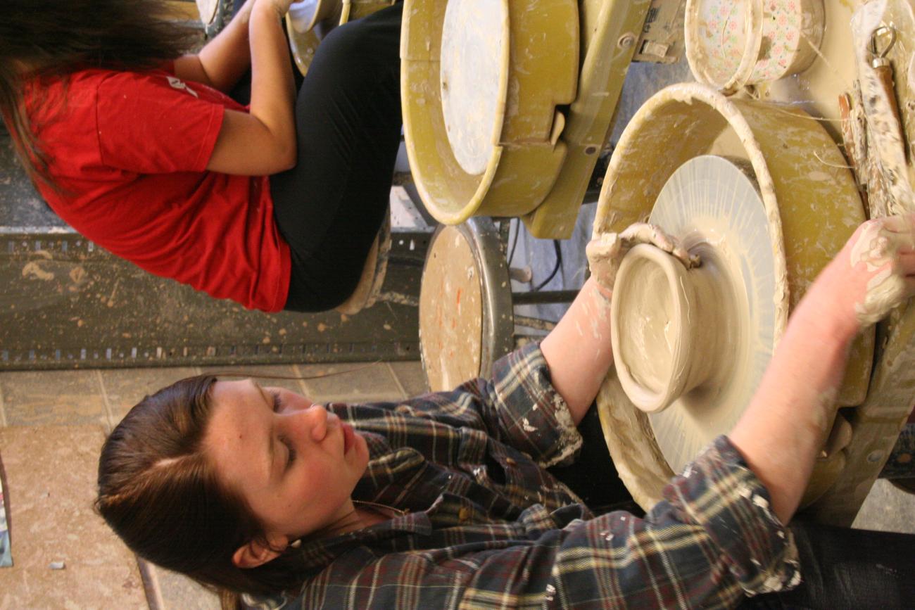 A student looks down calmly as they reach for a tool while holding a mid-progress bowl on a potter's wheel