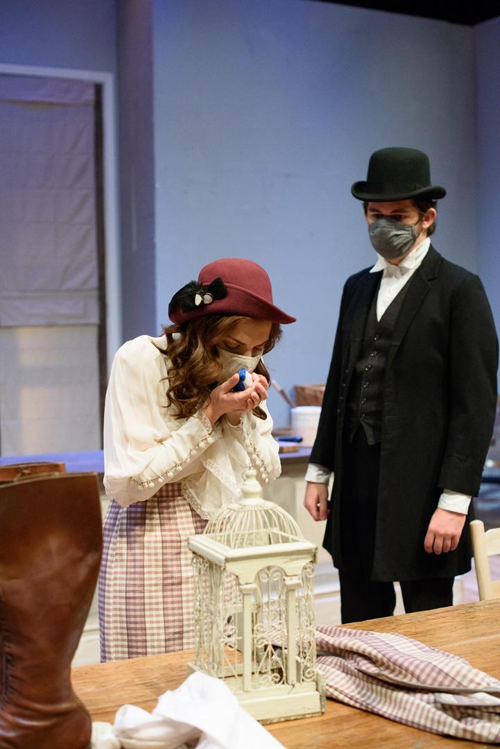 Actor in dress holds a bluebird out of its cage and whispers to it while another in a top hat looks on.