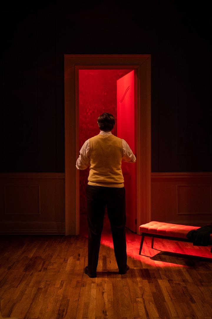 Actor in yellow vest and slacks is framed by a tall doorway they look into with red light spilling out of it.
