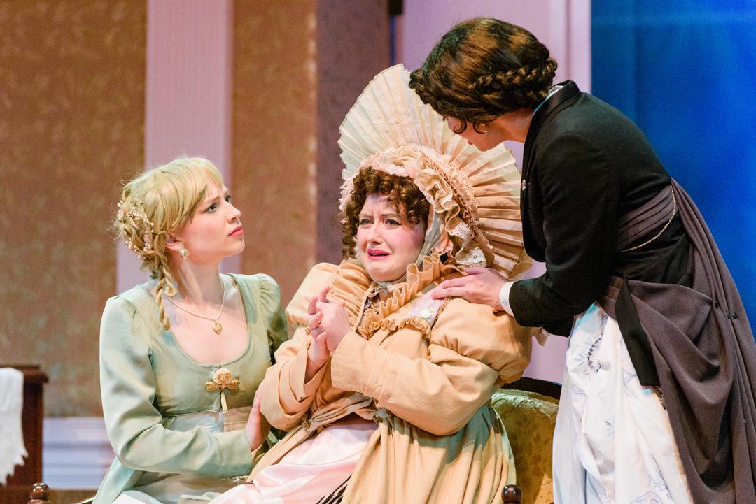 Mrs Bennet wearing a frilly, loud yellow outfit worries as two daughters comfort them.