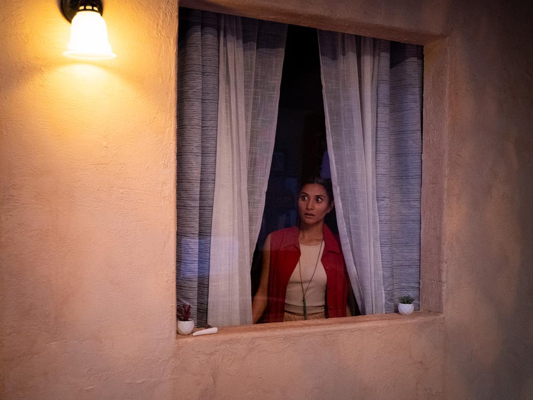 Actor looks shocked peering out of sheer curtains from a window as an outdoor light highlights the stucco of the wall.