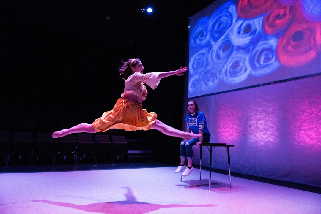 Dancer wearing a flowy orange skirt leaps as another enjoys sitting on a school desk with projected roses on the screen behind.