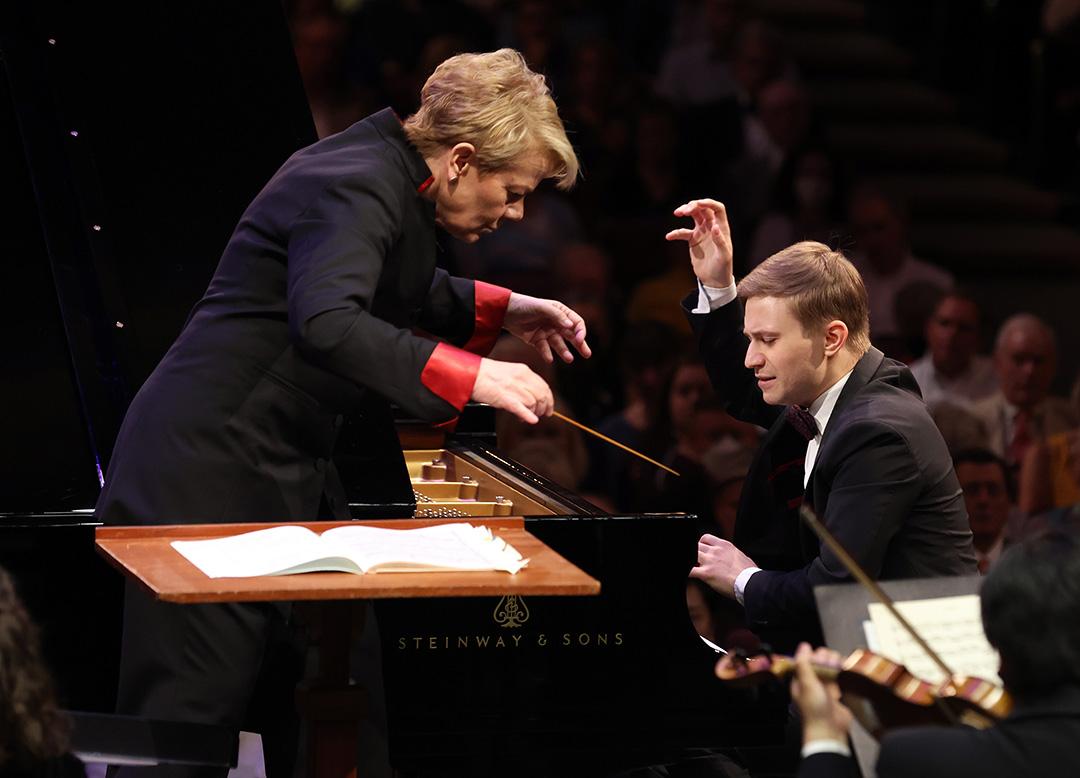 Dmytro looks down with one hand above piano keys and the other in the air like a claw, as a conductor leans over him watching the keys. A violinist plays in the foreground and a large audience watches