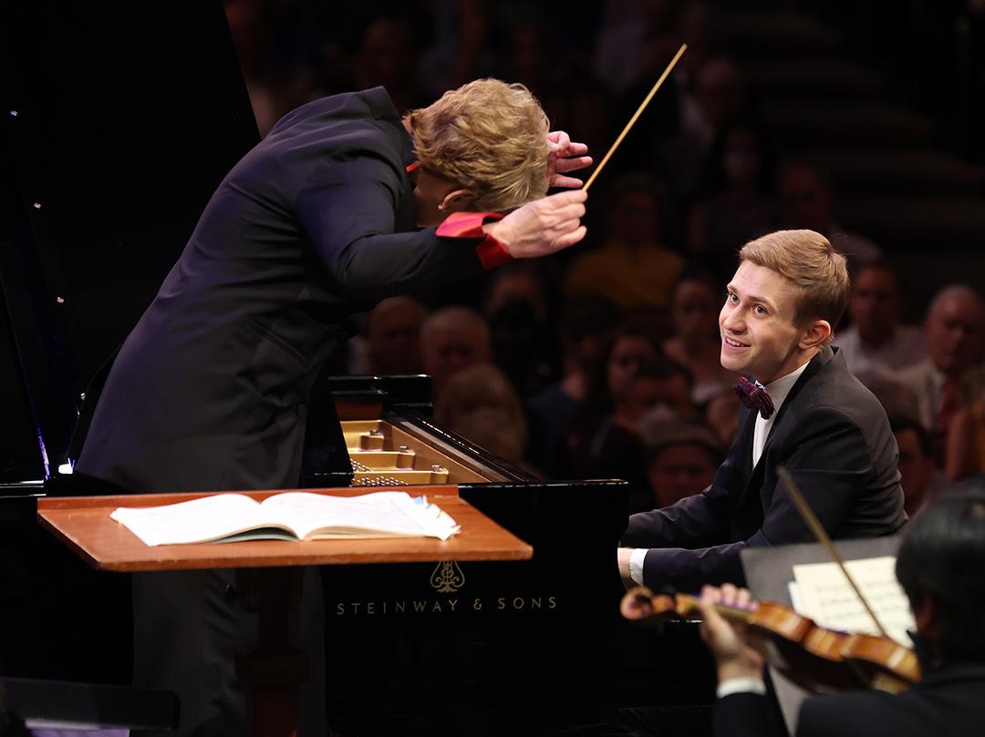 Dmytro smiles at the conductor while playing piano in front of a large audience. A violinist plays off to the side.