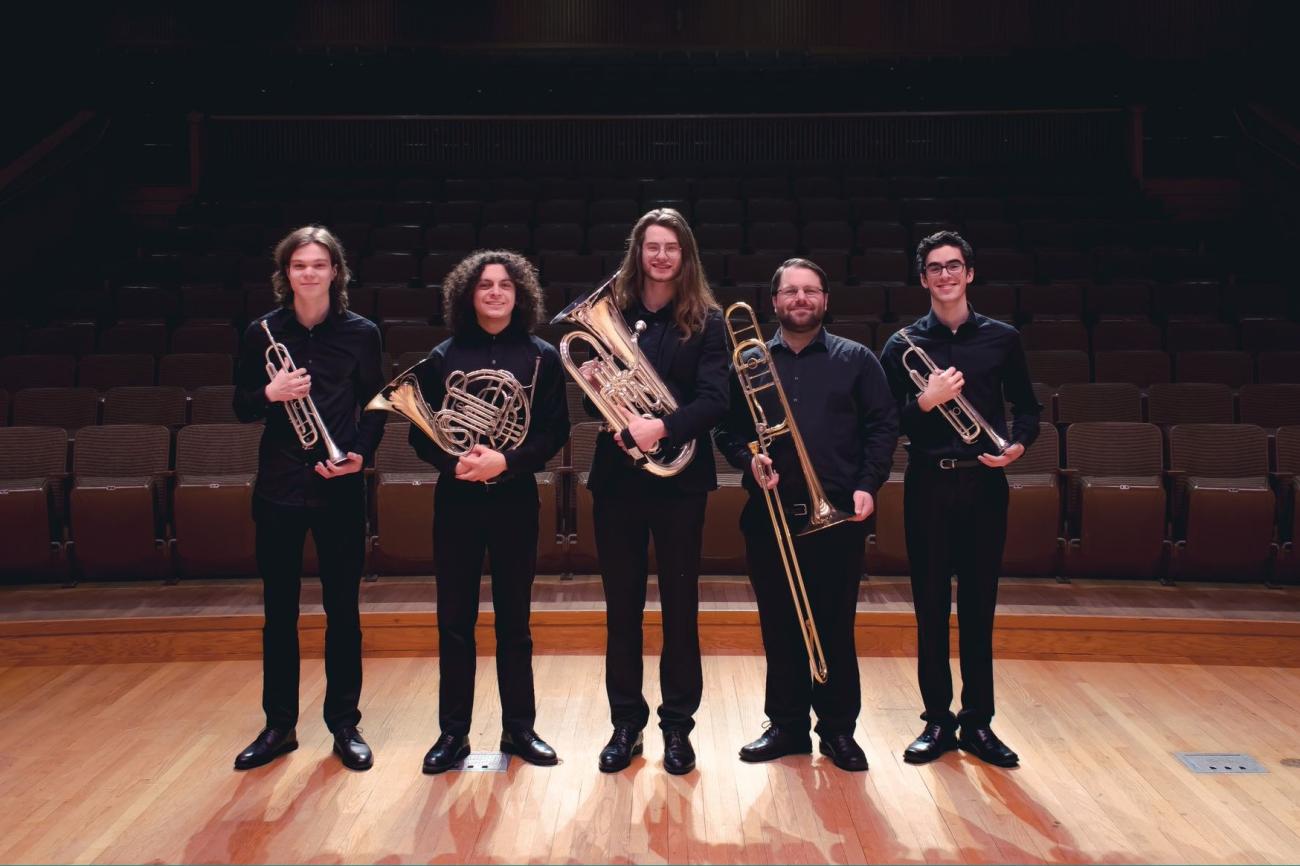 Five people stand side by side in formal attire, smiling and holding brass instruments