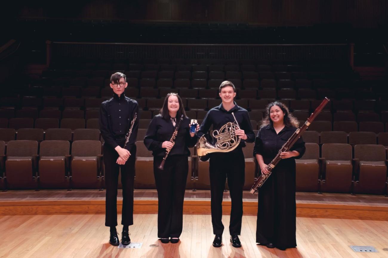 Four people stand side by side in formal attire, smiling and holding wind instruments