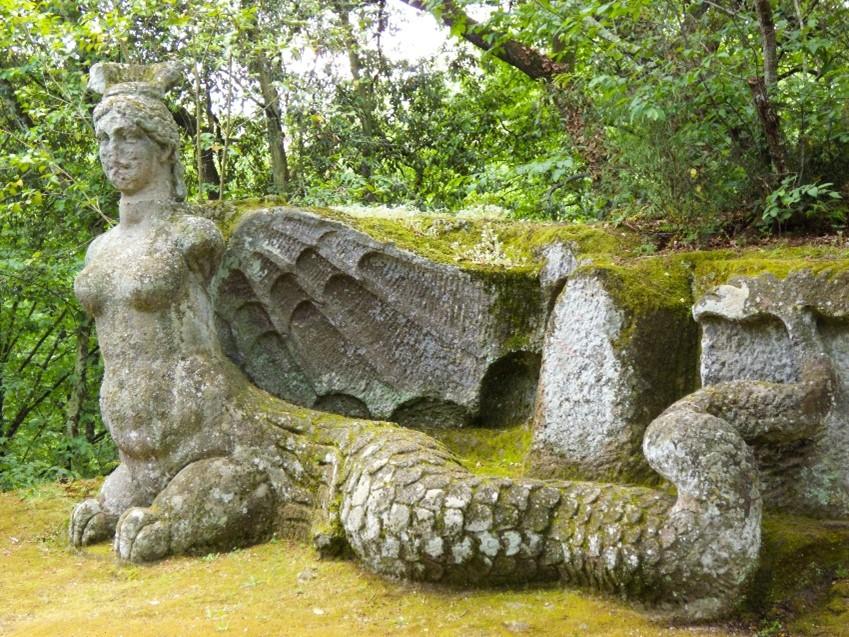 moss covered stone sculpture in the woods: limbless, winged woman with a long snake tail