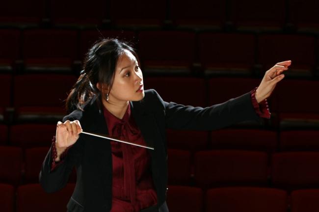 A person in formal attire focuses holds one hand out with a focused gaze in that direction, and the other hand holds a conductor's wand