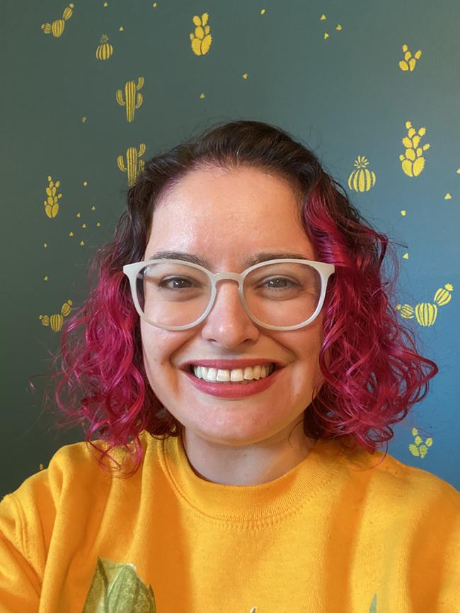 Ana María from the shoulders up with a large smile in front of a wall with little cactuses painted on it. Ana María has short, curly hair that fades from dark to bright pink, large round glasses, and a yellow sweatshirt