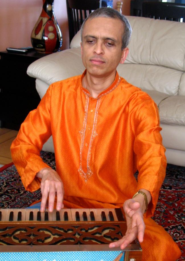 Mohan Bhide sitting on the floor in a bright orange traditional Indian suit, playing harmonium
