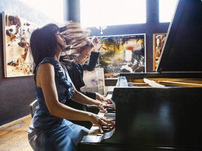 Anderson and Roe actively playing piano together. Anderson's left arm is up by his head. Roe's hair flies as her head tilts back with her eyes closed.