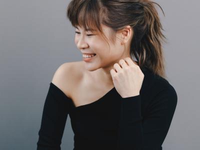 Rachel Cheung sitting cross legged, leaning forward mid-giggle with her face turned to the side and one hand up near her neck as if it was supporting her chin before she giggled
