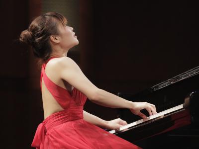 View from floor level of Rachel Cheung in a bright red, flowy formal gown, playing a Steinway piano on a stage. She is sitting with back straight and looking up with reverent emotion as she plays.
