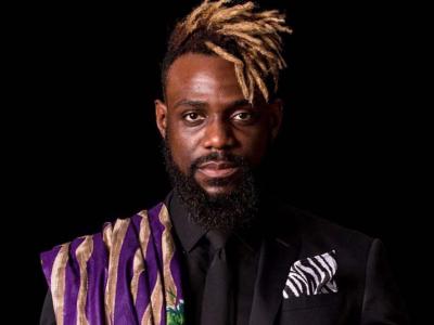 a black person with a beard, a patterned purple shawl over one shoulder, black suit, zebra striped kerchief in pocket, a shock of blond dredlocks spinging from the top of their head