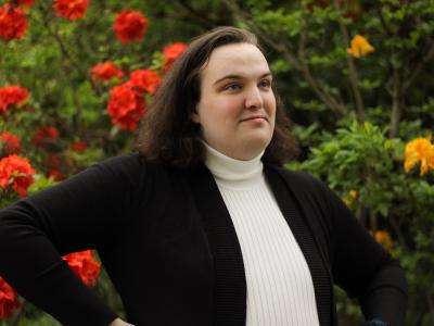 a person in a white turtleneck and black blazer stands in front of flowering shrubs
