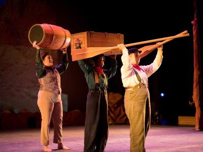 three actors holding a barrel, a crate, and a paddle over their respective heads