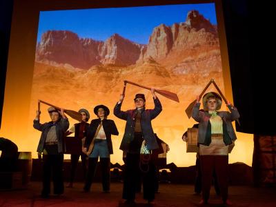 actors on stage holding broken boat paddles aloft. Behind is a projection of a rocky canyon wall.