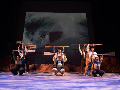 actors in a formation hold paddles over their heads as if descending rapids on a river