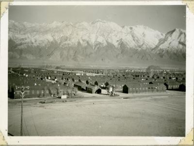 An aged black and white snapshot of ranks of barracks buildings against steep, aired snowcapped mountains.
