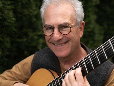 a smiling gray-haired person in glasses holds a guitar ready to play