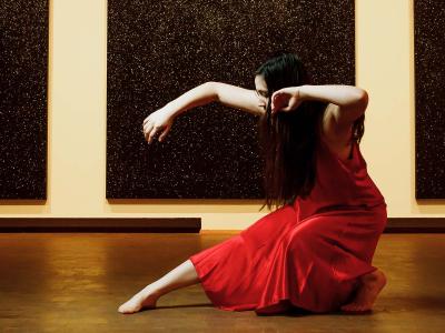 dancer in red leans back squatting low leg extended hair covers face. A black painting hangs behind.