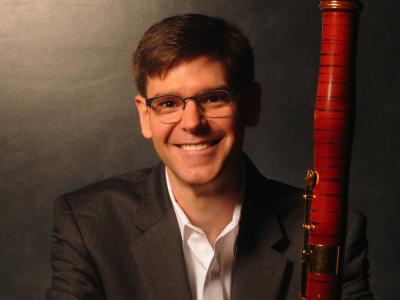 a person in a blazer and collared shirt wearing glasses smiles and cradles a bassoon