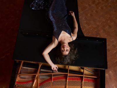 upside-down view from overhead: Inna Faliks in a dark sequined dress, laying on top of a steinway piano, smiling